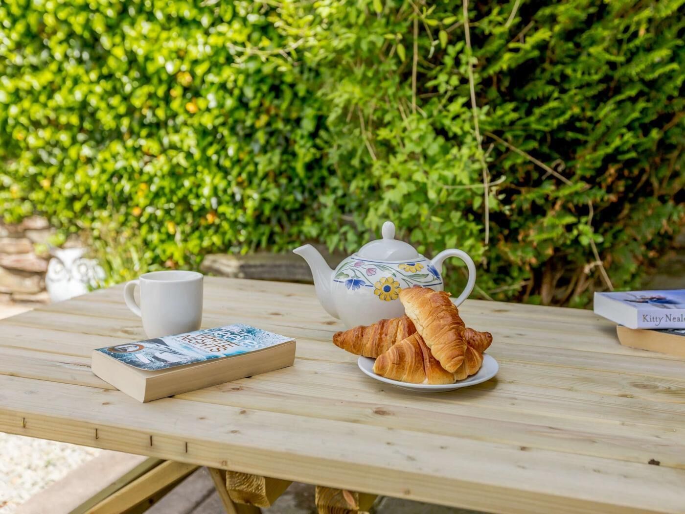 A wooden table with a teapot, a cup, a book, and a croissant.