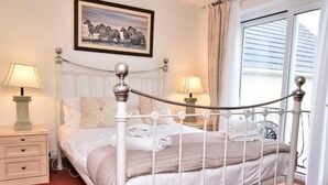 Ein Schlafzimmer mit weißem Metallbettgestell, einem Nachttisch mit Lampe, einem gerahmten Bild an der Wand und einem Fenster mit Vorhängen.