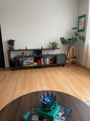 Ein Zimmer mit Holzboden, einem Bücherregal mit diversen Gegenständen, einem kleinen Tisch mit einem Dekorationsobjekt und einem Topfpflanzen-Ständer.