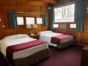 A room with two beds, wooden walls, a window with curtains, and a lamp on a nightstand.