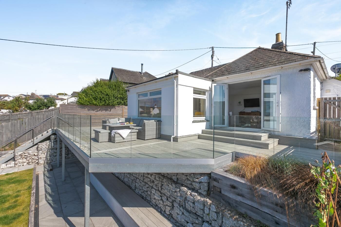 A modern house with a glass deck, outdoor seating, and a stone retaining wall.