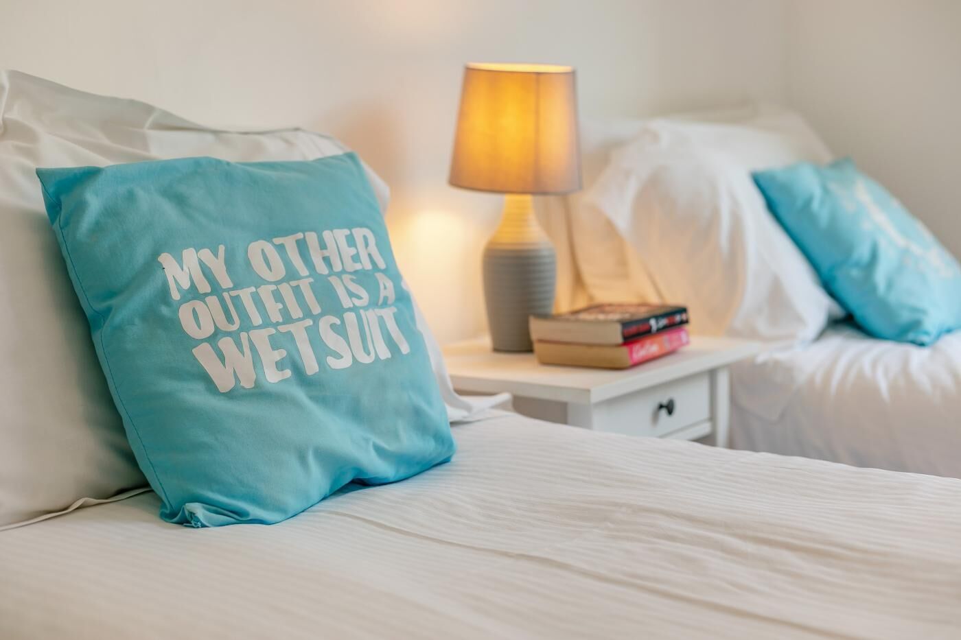 A bed with a pillow that reads 'MY OTHER OUTFIT IS A WETSUIT' and a bedside lamp.
