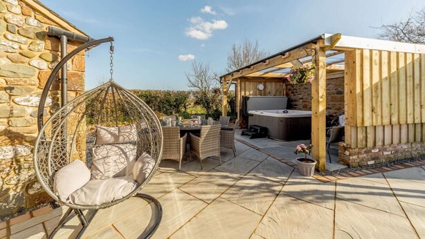 En terrasse med stenvæg, hængekøjsstol, et bord med stole og et spabad under et træpergola.