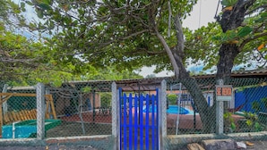 Un espace clos, délimité par une clôture, avec un portail bleu, une piscine et un panneau indiquant « À LOUER ».