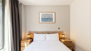 A neatly made bed with white linens, two bedside lamps, a framed picture on the wall, and a window with curtains. - Bright Lambeth Apartment by Waterloo (London)