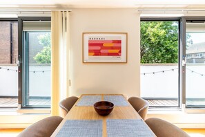 Un comedor con mesa, sillas, una obra de arte enmarco y ventanas amplias.