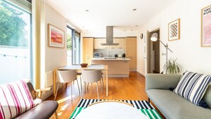 A modern living room with a sofa, a dining area with chairs, a kitchen with a central island, and a large window providing natural light. - Bright Lambeth Apartment by Waterloo (London)