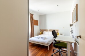 Ein Schlafzimmer mit einem Bett, einem Schreibtisch, einem Sessel, einer Lampe und einem Fenster mit Vorhängen.