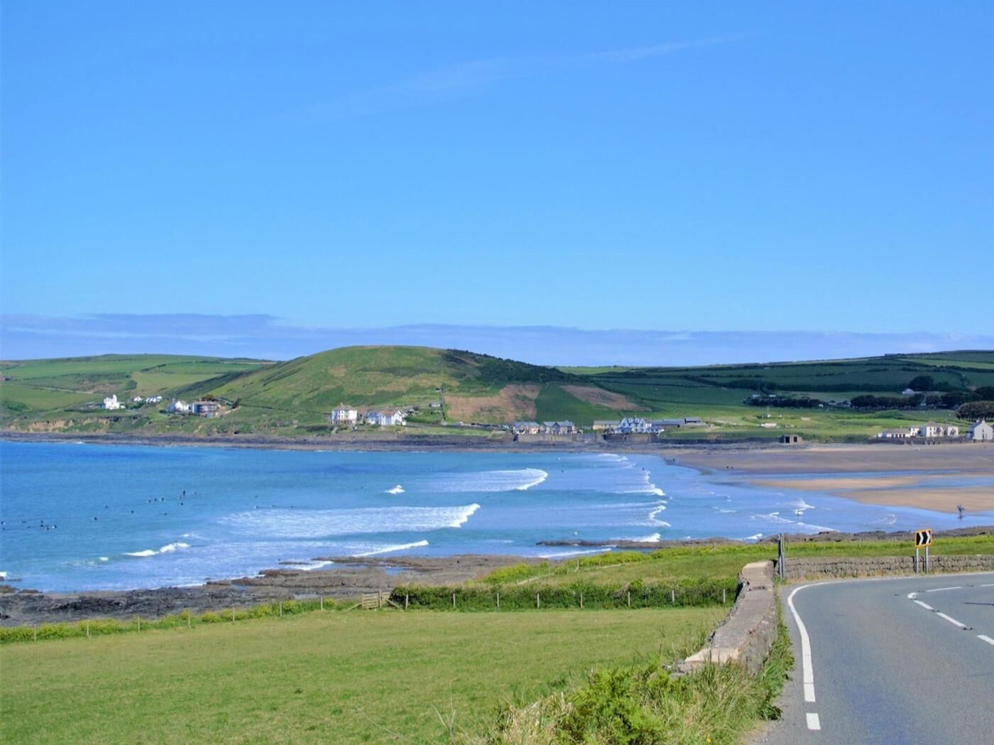 A coastal road with a view of the sea, grassy fields, and scattered houses.