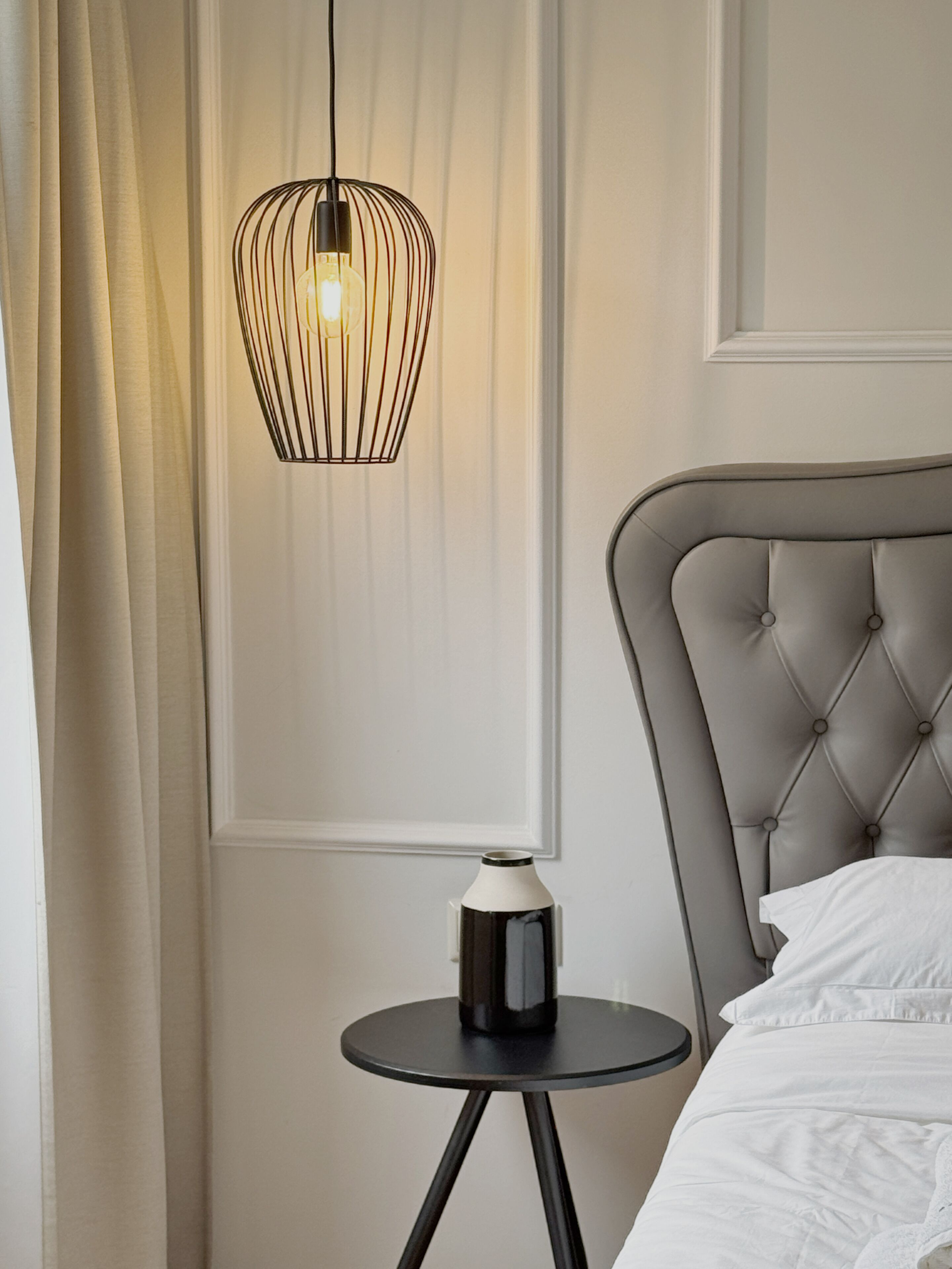 Une chambre à coucher avec une tête de lit capitonnée, une table de chevet avec une lampe noire et une fenêtre avec des rideaux.