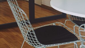 A modern interior with a round table and wireframe chairs, wooden flooring, and a glass partition with a black frame.