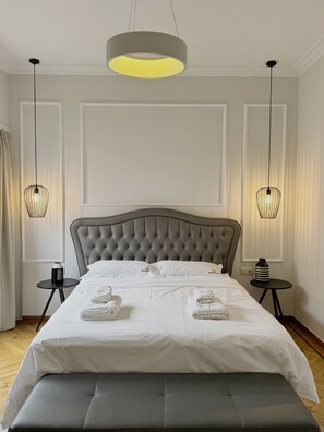 Quarto moderno com cabeceira estofada, cama grande com roupa de cama branca, duas mesas de cabeceira com abajures e um banco no pé da cama.