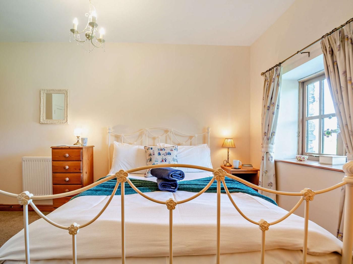 A bedroom with a bed, a dresser, a nightstand, a lamp, a window with curtains, and a chandelier.