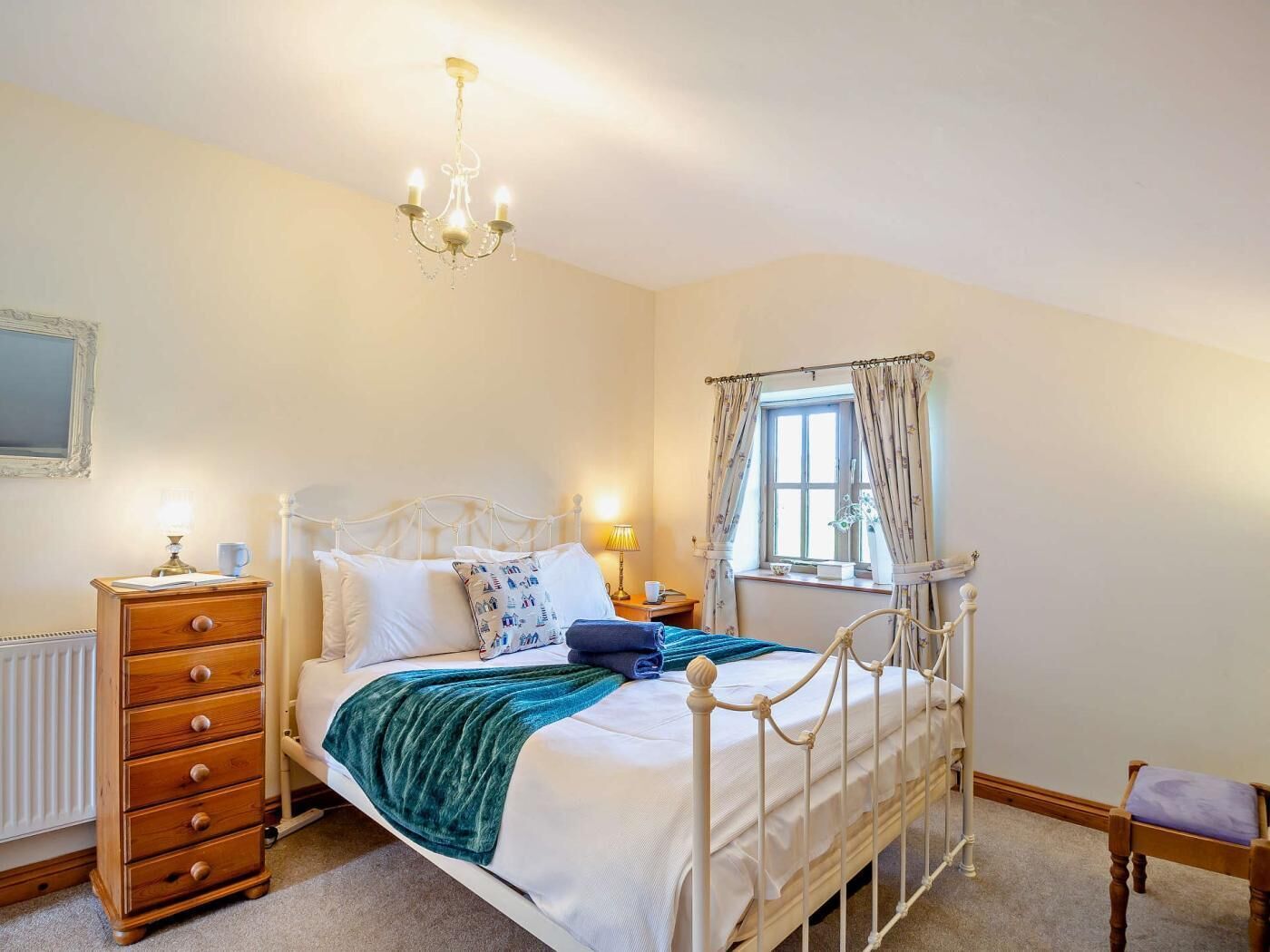 A bedroom with a bed, dresser, nightstand, lamp, window with curtains, and a chandelier.