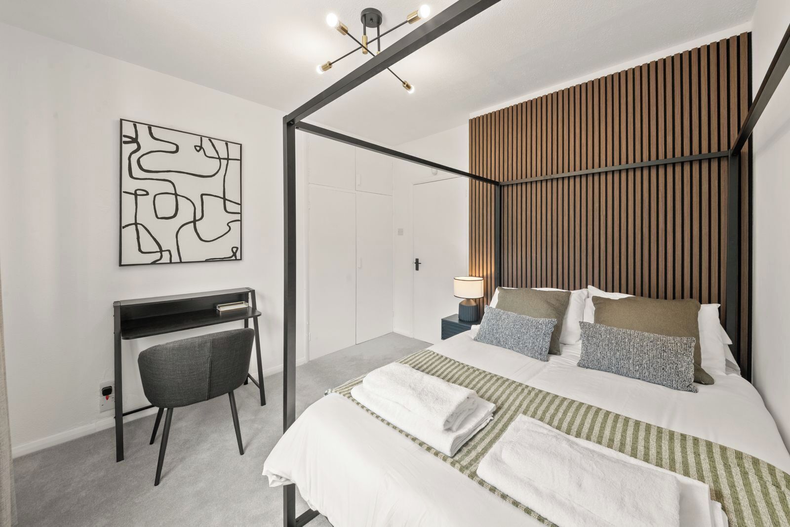 A modern bedroom with a bed, bedside table, lamp, desk, chair, and abstract artwork.