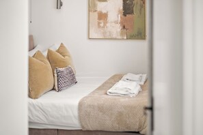 A neatly made bed with a white comforter, beige pillows, and a striped pillow. A folded towel is placed on a beige blanket on the bed. - Stylish home I sleeps 8 I free parking (Virginia Water)