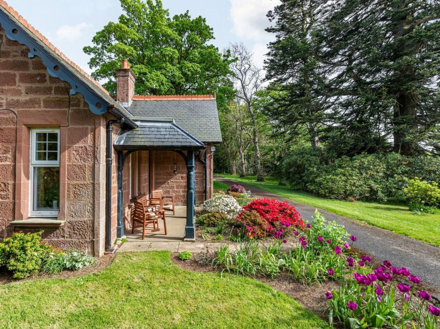 A stone building with a covered porch, a brick chimney, and a garden with flowers and shrubs.