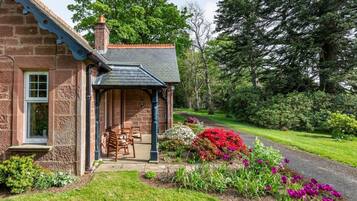 A stone building with a covered porch, a brick chimney, and a garden with flowers and shrubs.