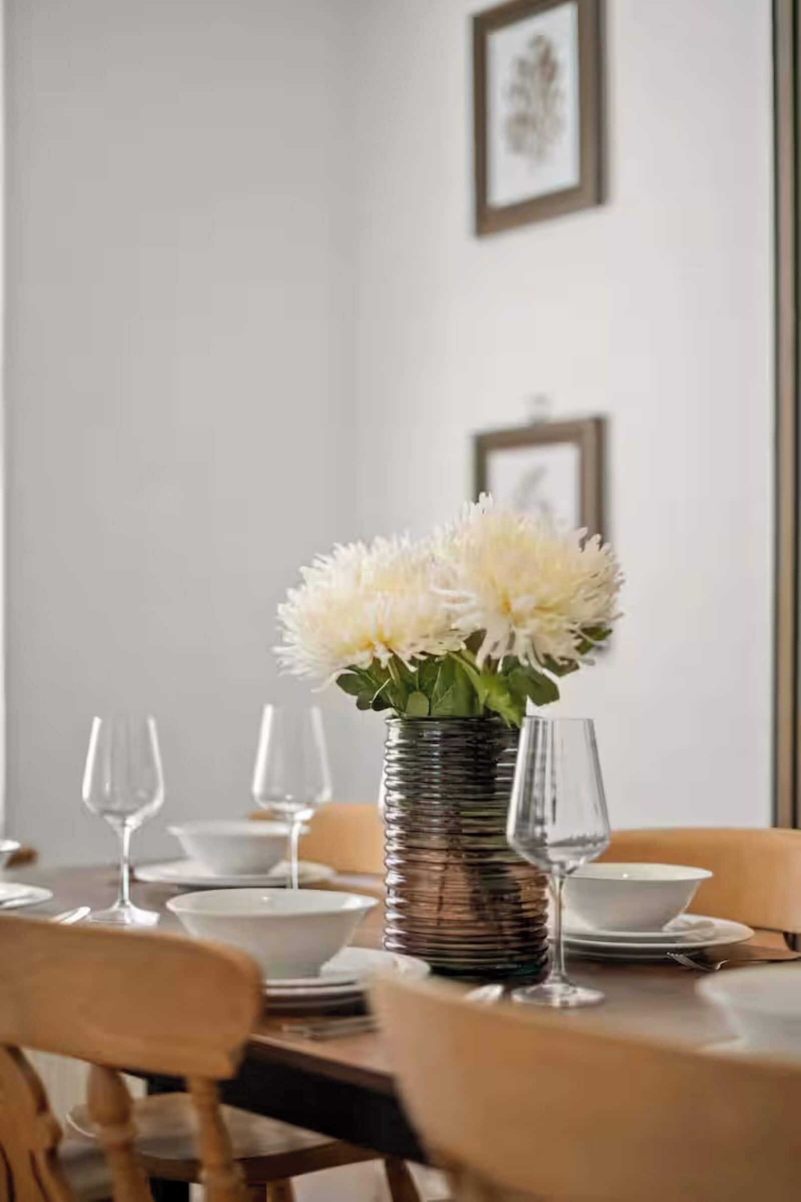 Una mesa de comedor dispuesta con platos, fuentes y copas de vino, con un florero en el centro.