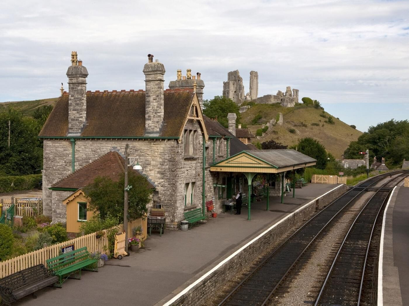 A stone building with a green roof, a covered waiting area, and a railway track.