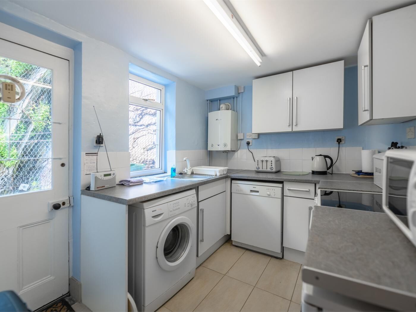 A compact kitchen with a washing machine, dishwasher, and a window with a view of greenery.
