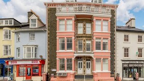 A multi-story building with a prominent pink facade, a central entrance with double doors, and a balcony with ornate railings. - 2 Bed in Aberdyfi (oc-dy039) (Aberdovey)
