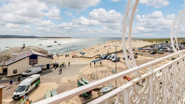 A beach view with a parking area, a building with a flag, and a white decorative railing. - 2 Bed in Aberdyfi (oc-dy039) (Aberdovey)