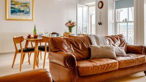 A living room with a brown leather sofa, wooden chairs, a dining table with a vase of flowers, and a painting on the wall. - 2 Bed in Aberdyfi (oc-dy039) (Aberdovey)