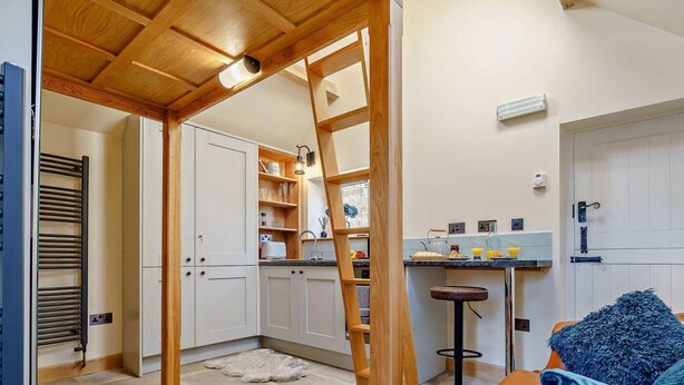A cozy interior with a wooden ceiling, a kitchen area with cabinets and a sink, a stone-tiled floor, and a leather sofa with a blue pillow. - 1 Bed in Whitby (oc-86298) (Castleton)