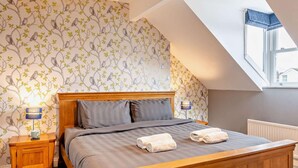Ein Schlafzimmer mit schrägen Wänden, einem Bett mit grauer Bettwäsche, zwei Nachttischlampen und einem Fenster mit Blick.
