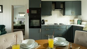 A modern kitchen with dark cabinets, a microwave, oven, and a gas stove. There is a dining table with plates and a glass of orange juice. - 2 Bed in Castle Douglas (oc-86449) (Auchencairn)