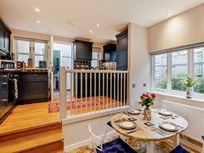 Une cuisine moderne avec des meubles de couleur sombre, un coin repas avec une table ronde dressée pour quatre personnes, et une vue sur un jardin par la fenêtre.