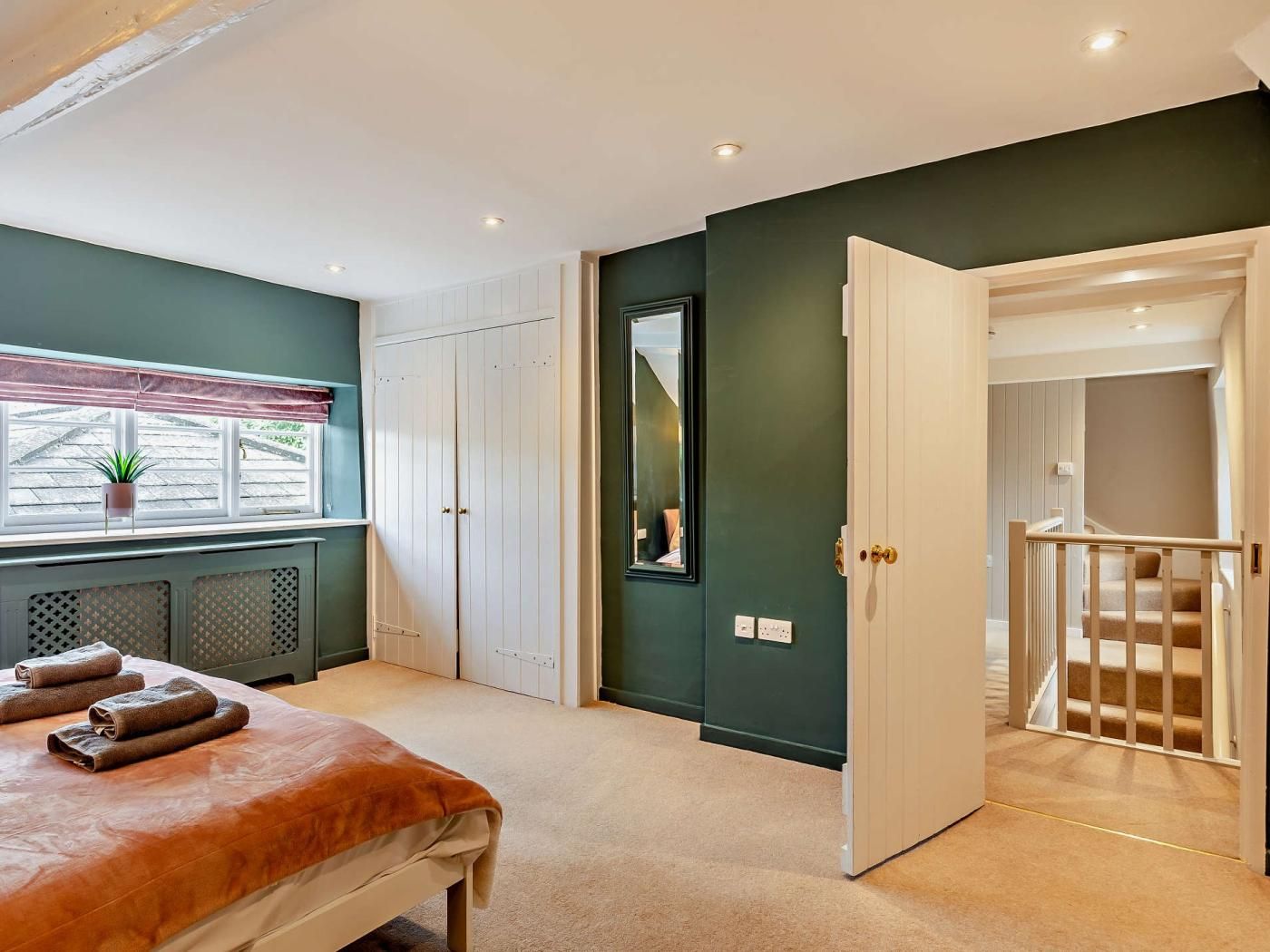 Une chambre à coucher avec un lit, une fenêtre ornée d’une plante, une armoire et une porte donnant sur un escalier.
