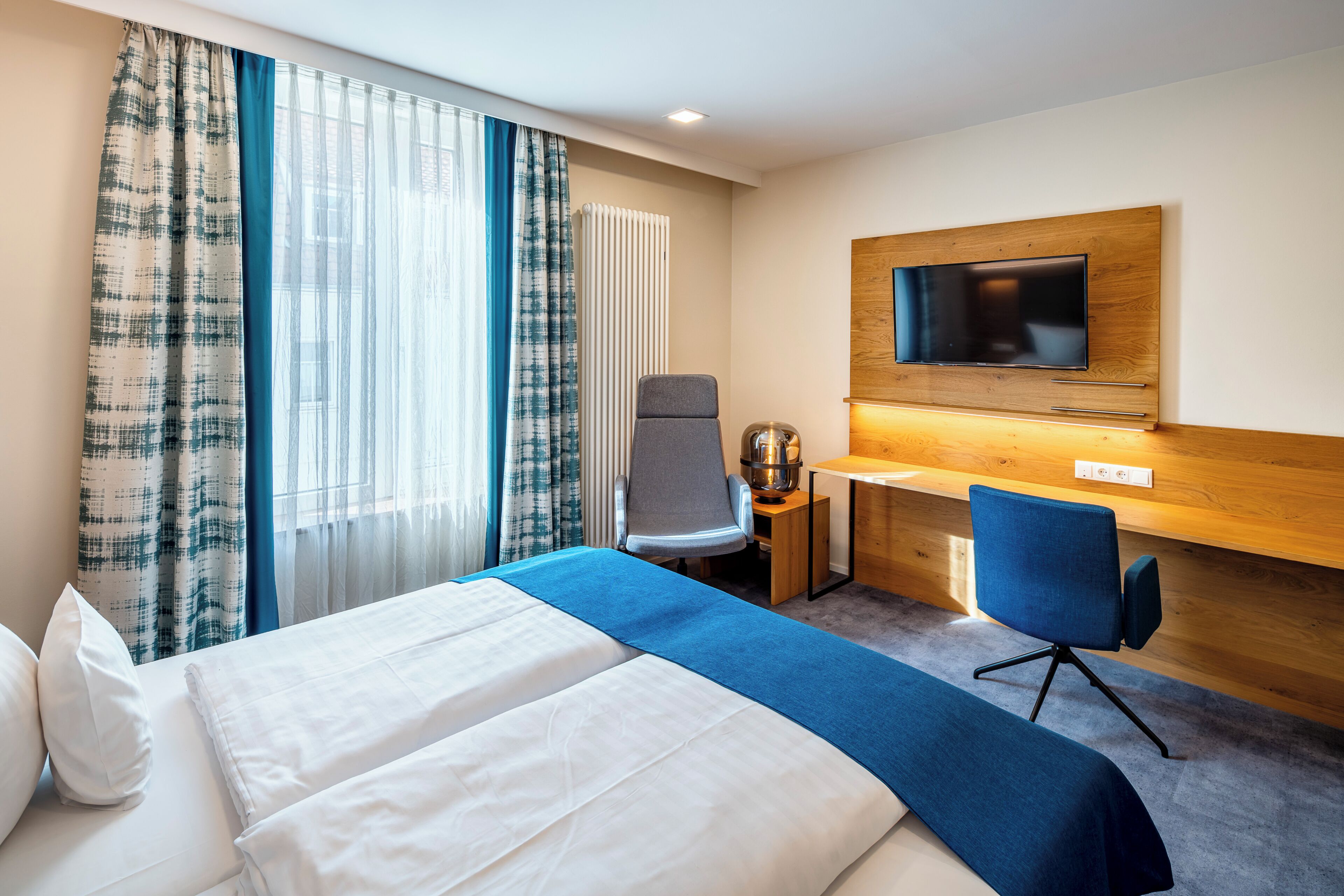 Una habitación de hotel con cama, dos sillas, escritorio, televisión y ventana con cortinas.