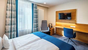 Una habitación de hotel con cama, dos sillas, escritorio, televisión y ventana con cortinas.