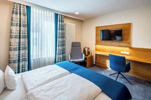 A hotel room with a bed, two chairs, a desk, a television, and a window with curtains.