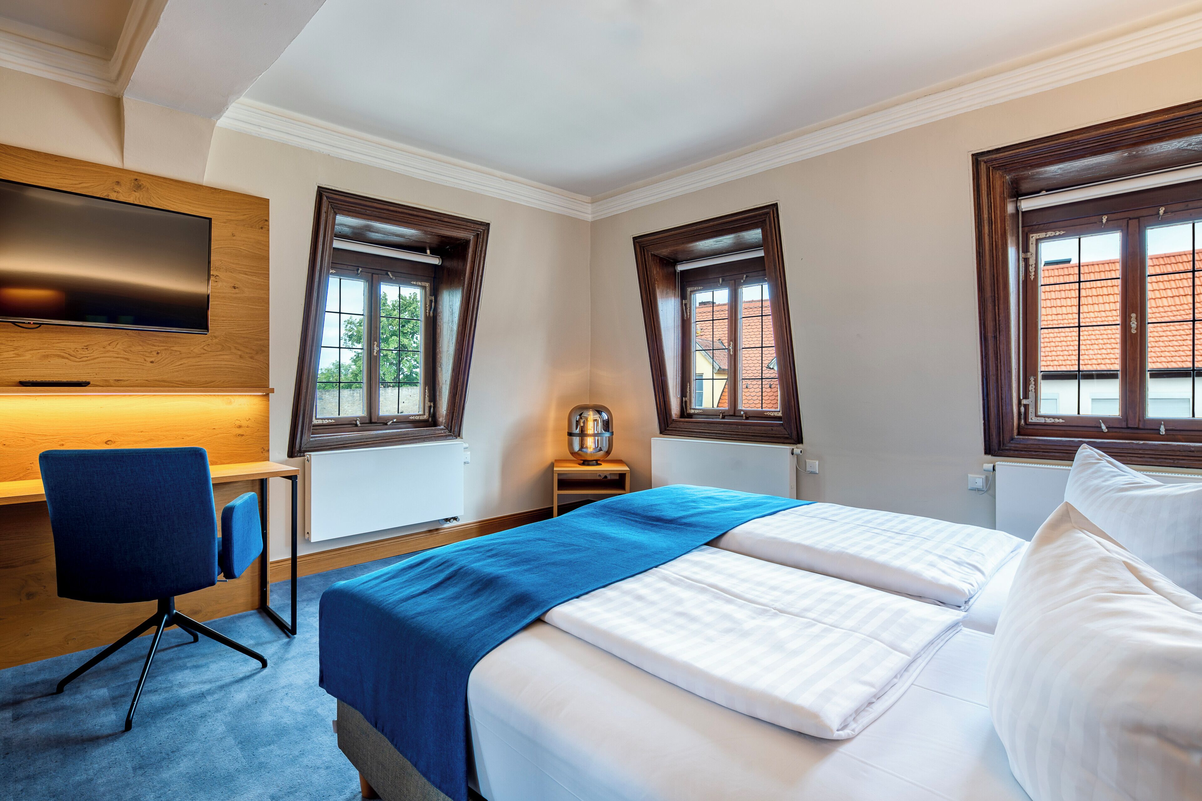 Quarto de hotel com cama, escrivaninha, cadeira, televisão e duas janelas.