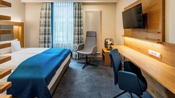 A hotel room with a bed, a desk, a chair, a television, and a radiator.