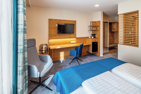 A hotel room with a bed, a desk with a chair, a TV, a wardrobe, and a small kitchenette.