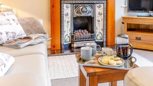 Un salon chaleureux avec une cheminée, une table basse avec des tasses et des biscuits, et un canapé avec un magazine.
