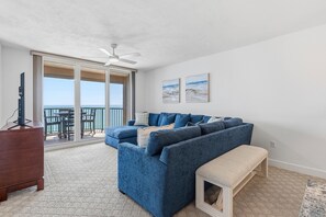 Ein Wohnzimmer mit blauem Sofa, einer Holzbank, einem Fernseher auf einem Schrank und einem Balkon mit Meerblick.