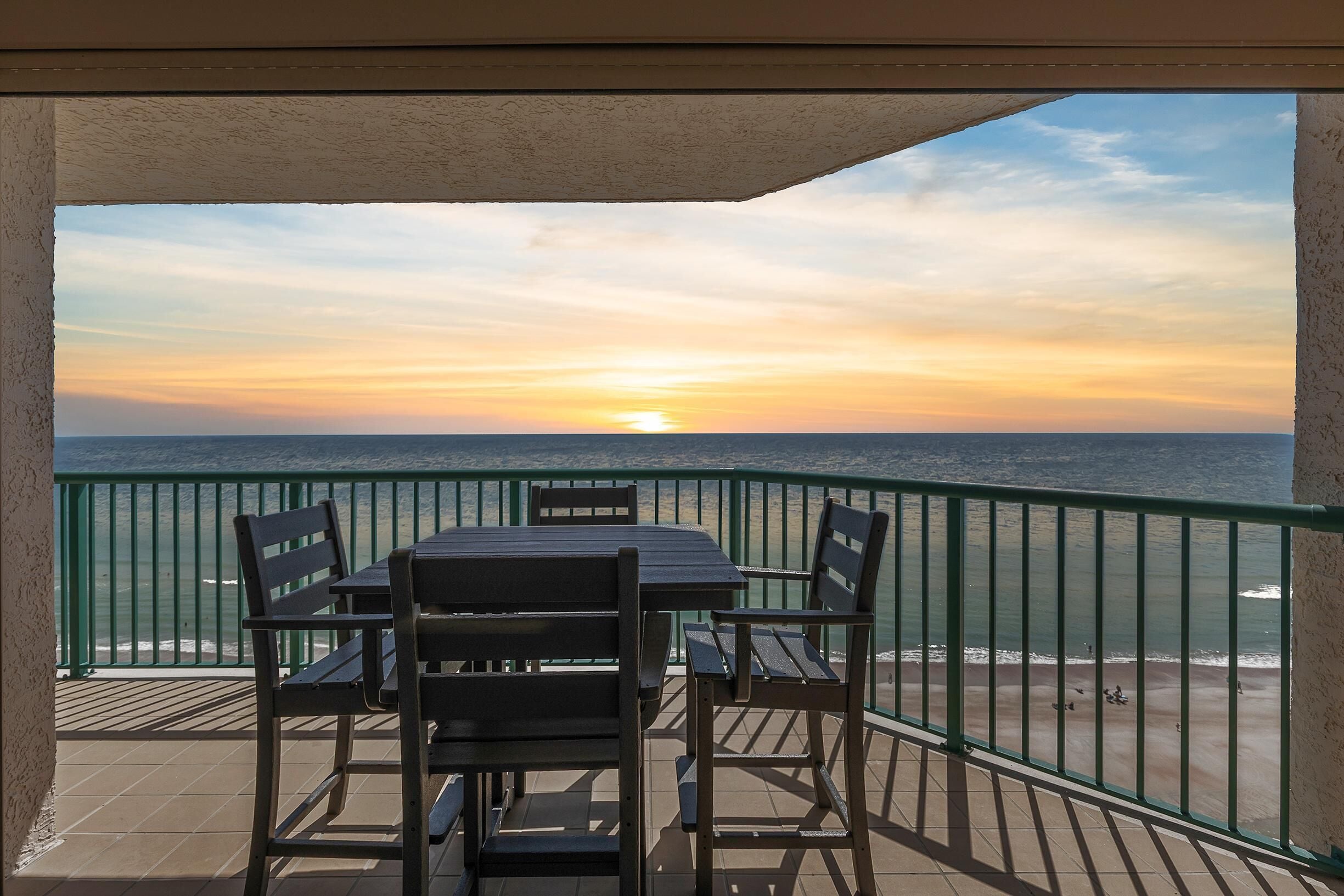 Ein Balkon mit Tisch und Stühlen, mit Blick auf einen Strand und das Meer bei Sonnenuntergang.