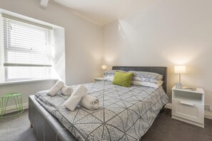 A bedroom with a bed, two bedside tables, a window with blinds, and a green stool. - 3 Bed in Tenby (oc-86574) (Tenby)