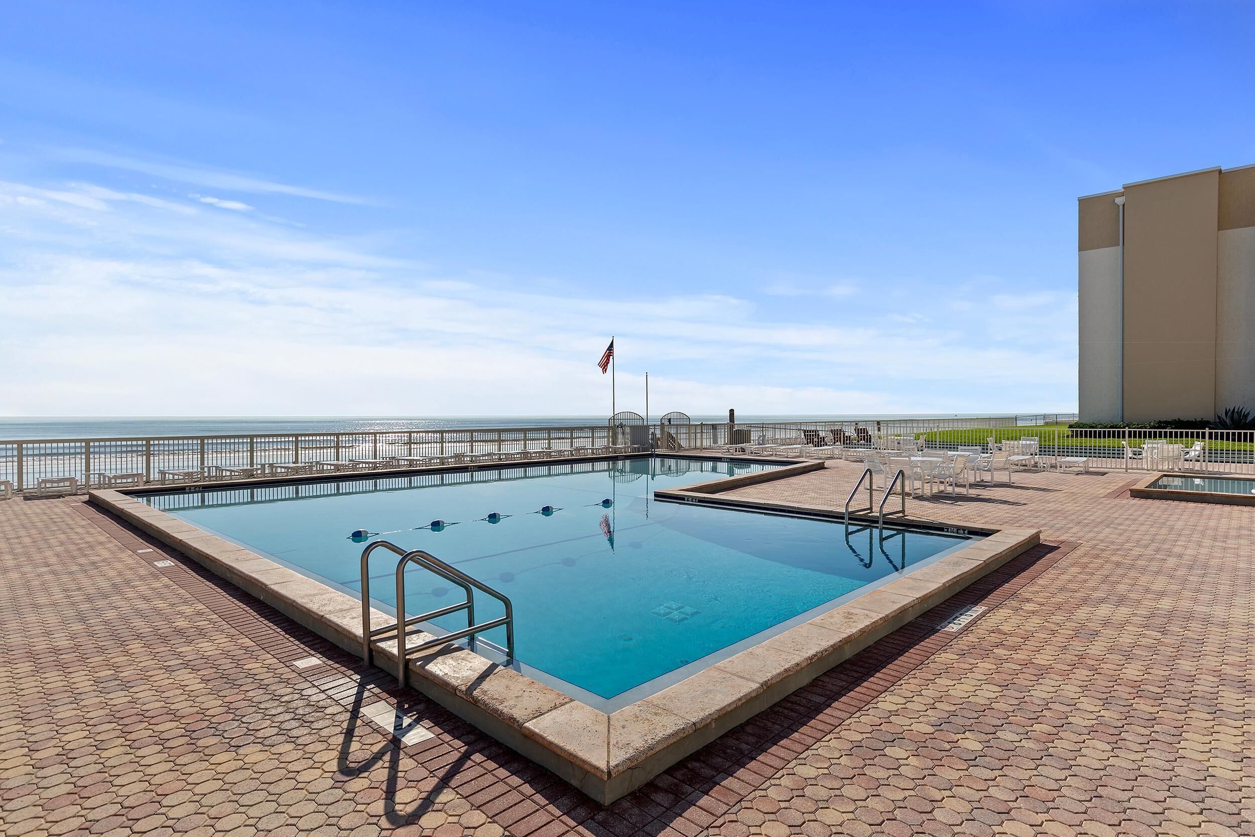 Uma piscina retangular com um deck branco, uma bandeira e vista para a praia.