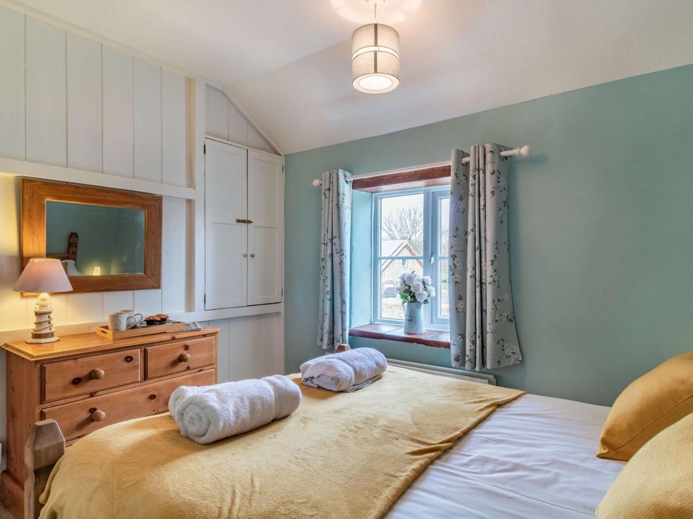 A bedroom with a bed, a dresser, a mirror, a window with curtains, and a lamp.