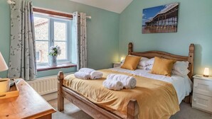 Een slaapkamer met een houten bed, een raam met gordijnen, een nachtkastje met een lamp, en een schilderij aan de muur.