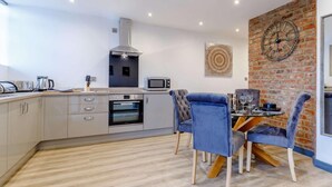 Uma cozinha moderna com forno embutido, micro-ondas e fogão a gás. Há uma mesa de jantar com cadeiras e uma parede de tijolos com um relógio.