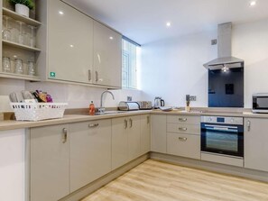 Cuina moderna amb forn integrat, tetera i torradora. Hi ha armaris, un rentapeus i una finestra amb persianes.
