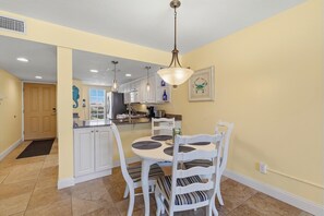 A dining area with a round table, white chairs, a hanging light fixture, and a kitchen in the background. - Point East 205 (New Smyrna Beach)