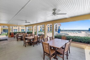 A covered outdoor dining area with tables and chairs, a ceiling fan, and a view of a pool and beach. - Point East 205 (New Smyrna Beach)